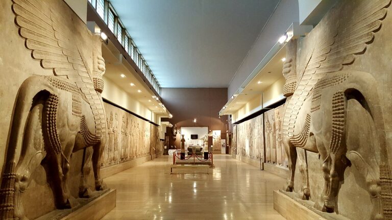 An impressive museum hall featuring large, winged bull statues flanking walls adorned with ancient reliefs, leading to an exhibition area in the distance.