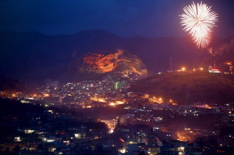 A nighttime view of a city with lights twinkling against a backdrop of mountains and a fireworks display lighting up the sky.