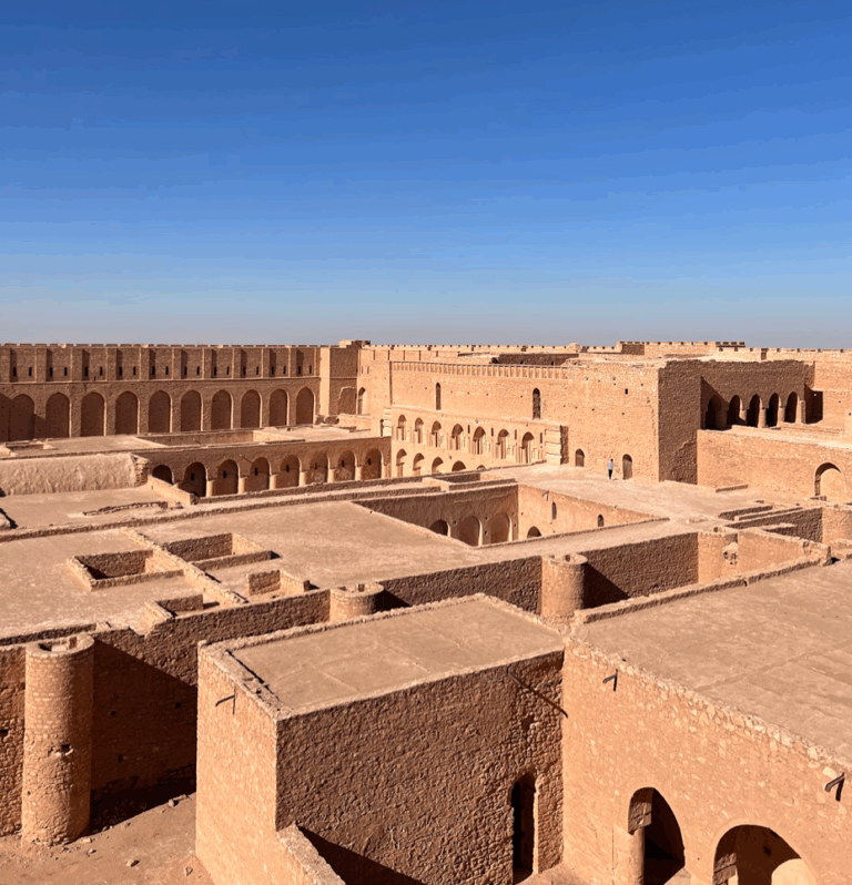 An aerial view showcases a vast, sandy-colored fortress with multiple levels, arches, and structures under a clear blue sky.