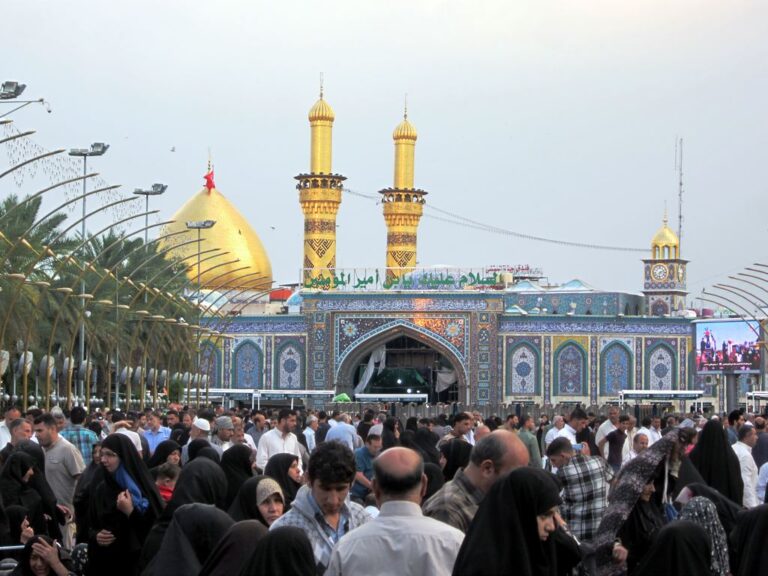 A large crowd gathers in front of a beautifully decorated mosque with golden domes and minarets.