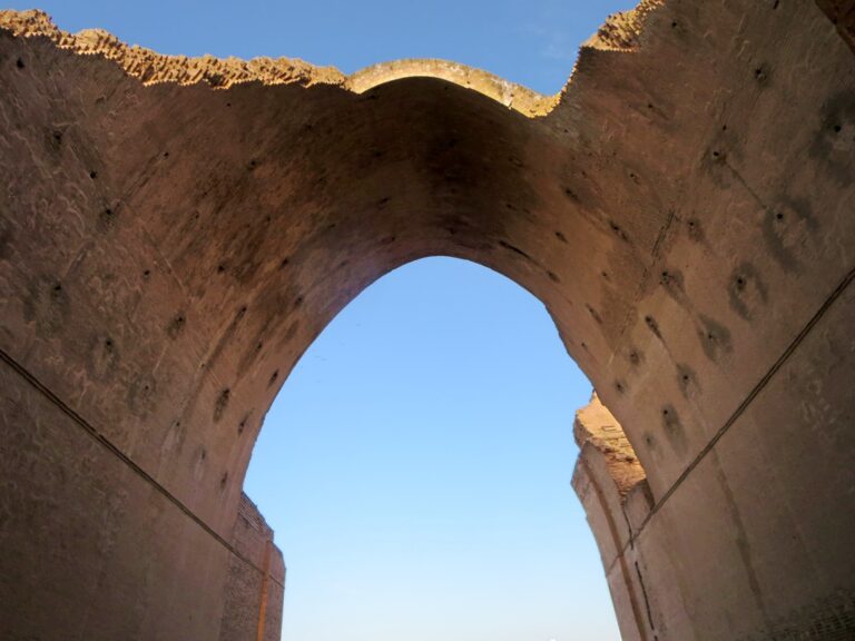 A low-angle shot captures the grandeur of an ancient archway or ruin, framing a clear blue sky with its weathered brickwork and crumbling edges.