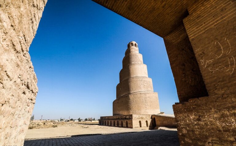The Great Mosque of Samarra, with its iconic spiral minaret, stands tall against a clear blue sky, framed by the ancient brickwork of a nearby archway.