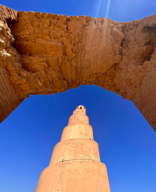 An upward view of the Great Mosque of Samarra through an arch, showing its spiral minaret against a clear blue sky.