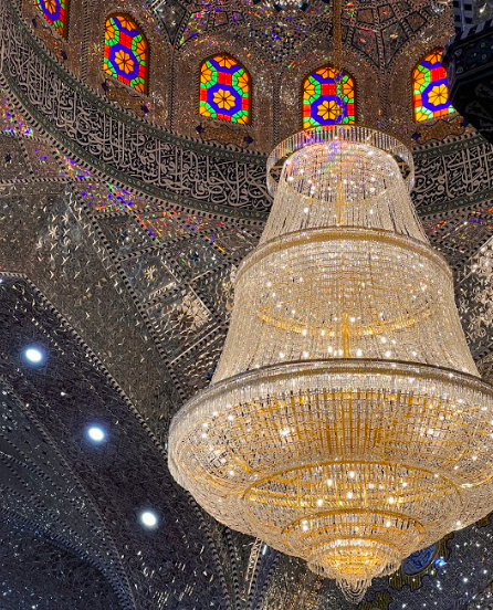 A large, ornate crystal chandelier hangs from the ceiling of a mosque, adorned with stained-glass windows and Arabic calligraphy.