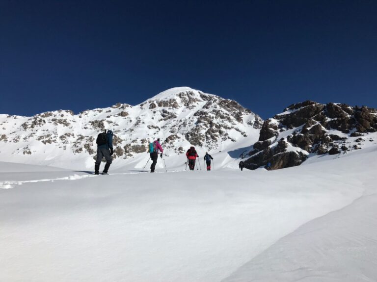 A group of hikers trek through a vast snow-covered landscape with towering mountains and a clear blue sky in the background.