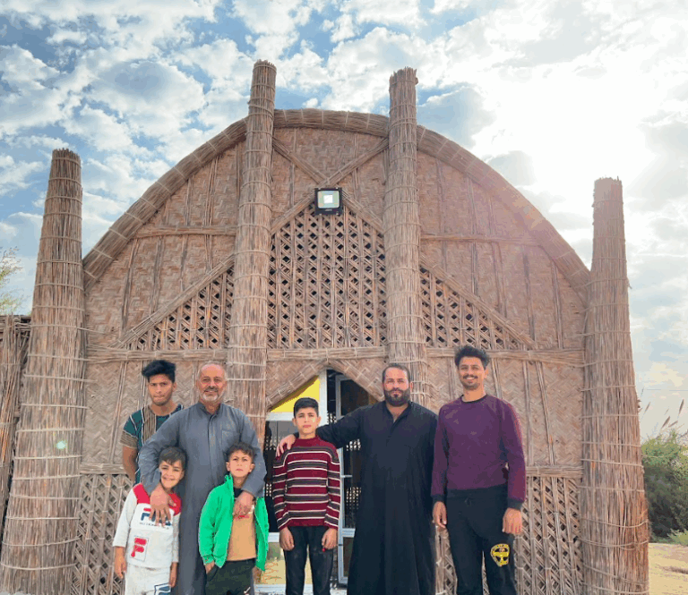 A group of seven people stands in front of a unique building constructed from natural materials, likely reeds or straw, resembling a mosque or a culturally significant structure.