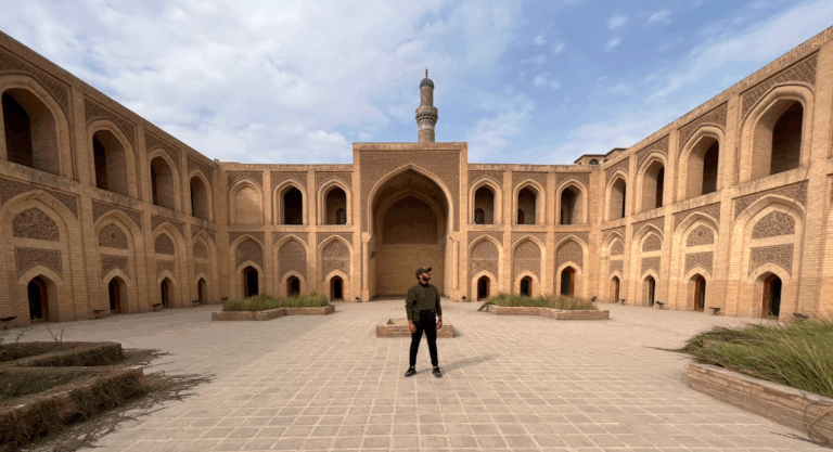 A man stands in the courtyard of the Abbasid Palace in Baghdad, Iraq. The palace features intricate brickwork, arched windows, and a minaret.