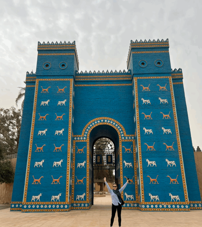 A woman stands in front of the Ishtar Gate, a massive blue structure adorned with animal figures. She has her arms raised in celebration.