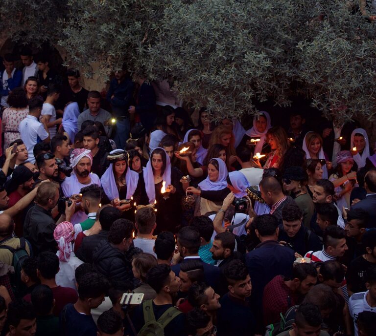 A crowd gathers with lit candles, many wearing traditional head coverings, under the branches of a tree, possibly during a religious or cultural event.
