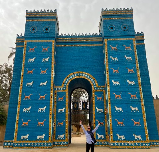 A smiling woman stands in front of a blue, ancient-looking gate decorated with alternating white and orange animals.
