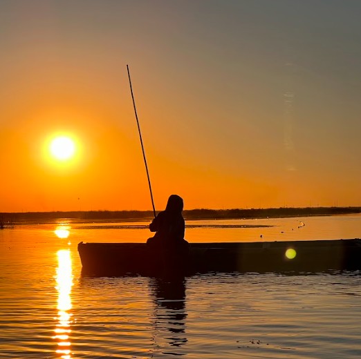 A silhouetted figure sits in a small boat on the water, holding a long pole, against a backdrop of a brilliant orange sunset.