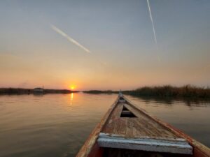 Day Tour Mesopotamian Marshes (Al-Ahwar) A boat navigates a serene river at sunset, with the sun reflecting on the water and contrails streaking the sky.