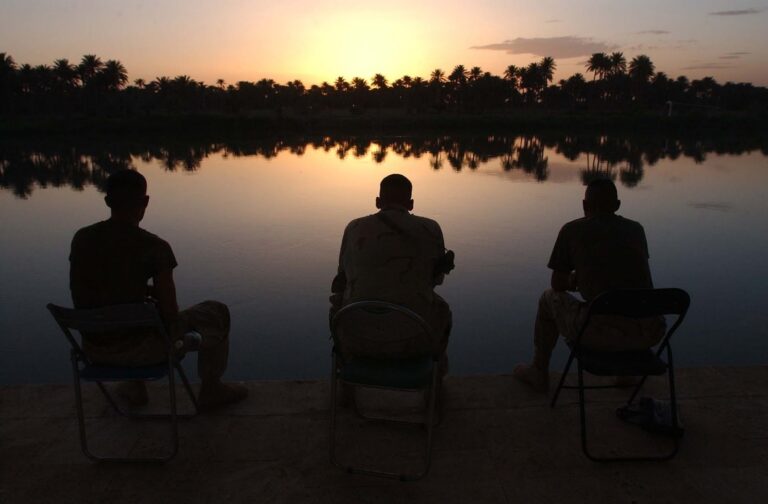 Three soldiers sit on chairs by a calm river at sunset, silhouetted against the vibrant sky and reflected trees.