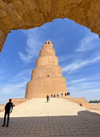 A striking spiral minaret, the Great Mosque of Samarra, framed by an arched entryway, with people visiting under a blue sky.