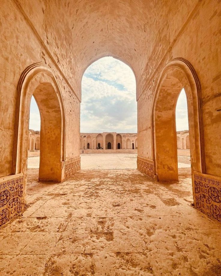 An arched opening leads to a large courtyard with more arches under a cloudy sky, all bathed in a warm, sandy tone.