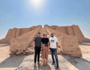 Day Tour from Nasiriyah – Ziggurat of Ur Three people stand smiling in front of the ancient Ziggurat of Ur under a bright, sunny sky.