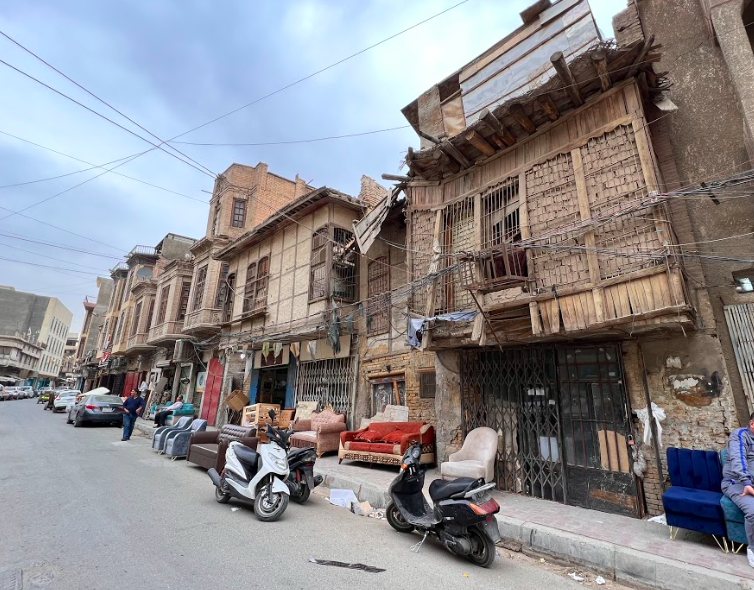 A street scene with old buildings and various items for sale on the sidewalk, including furniture and scooters.