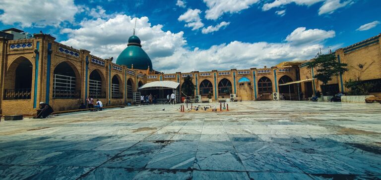 A wide shot of a tan building with arches and a turquoise dome, with people sitting and standing around in the courtyard on a partly cloudy day.