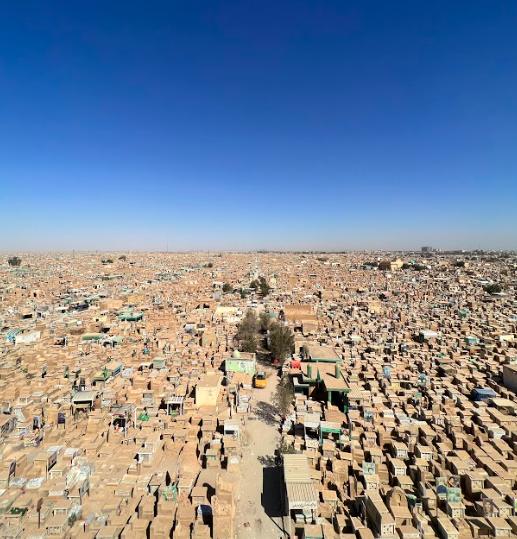 An aerial view shows an endless expanse of closely packed, weathered structures, likely grave markers, under a clear blue sky.