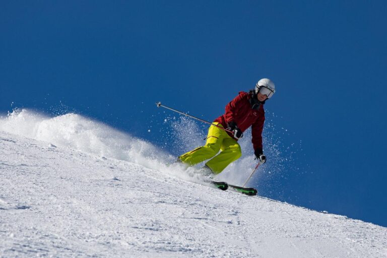 A skier in a red jacket and yellow pants carves down a snowy slope under a clear blue sky, kicking up snow as they turn.