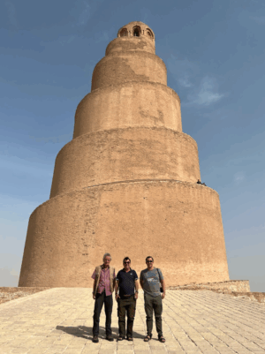 Day Tour Samarra Three men stand smiling in front of the Great Mosque of Samarra, a massive, spiraling, mud-brick minaret in Iraq, under a bright blue sky.