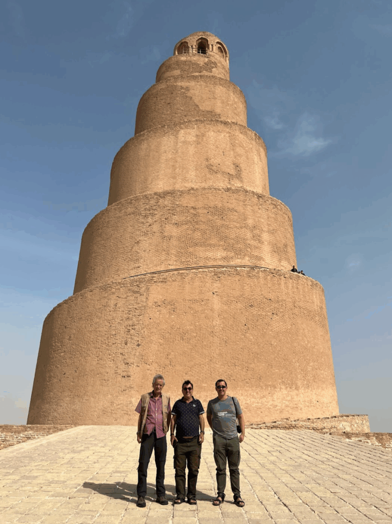 Three men stand smiling in front of the Great Mosque of Samarra, a massive, spiraling, mud-brick minaret in Iraq, under a bright blue sky.