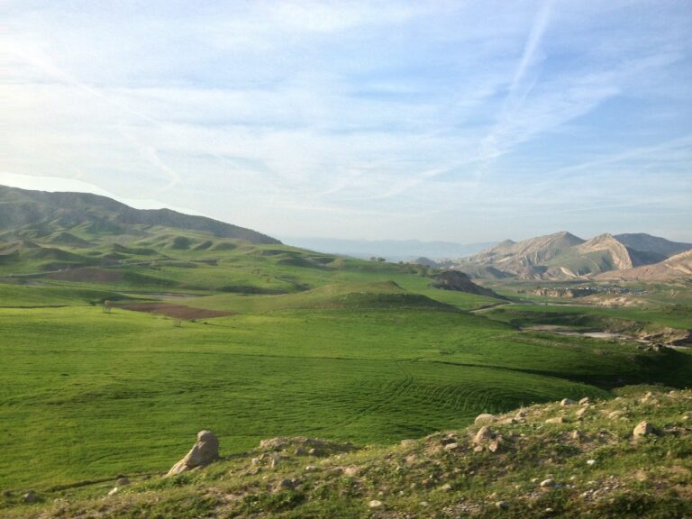 A vast, vibrant green landscape unfolds with rolling hills and distant mountains under a bright sky streaked with vapor trails.