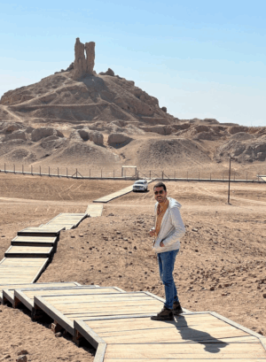 Day Tour Babylon from Baghdad A man stands on a wooden walkway leading to a mound of ruins, with a car parked nearby under a clear sky.