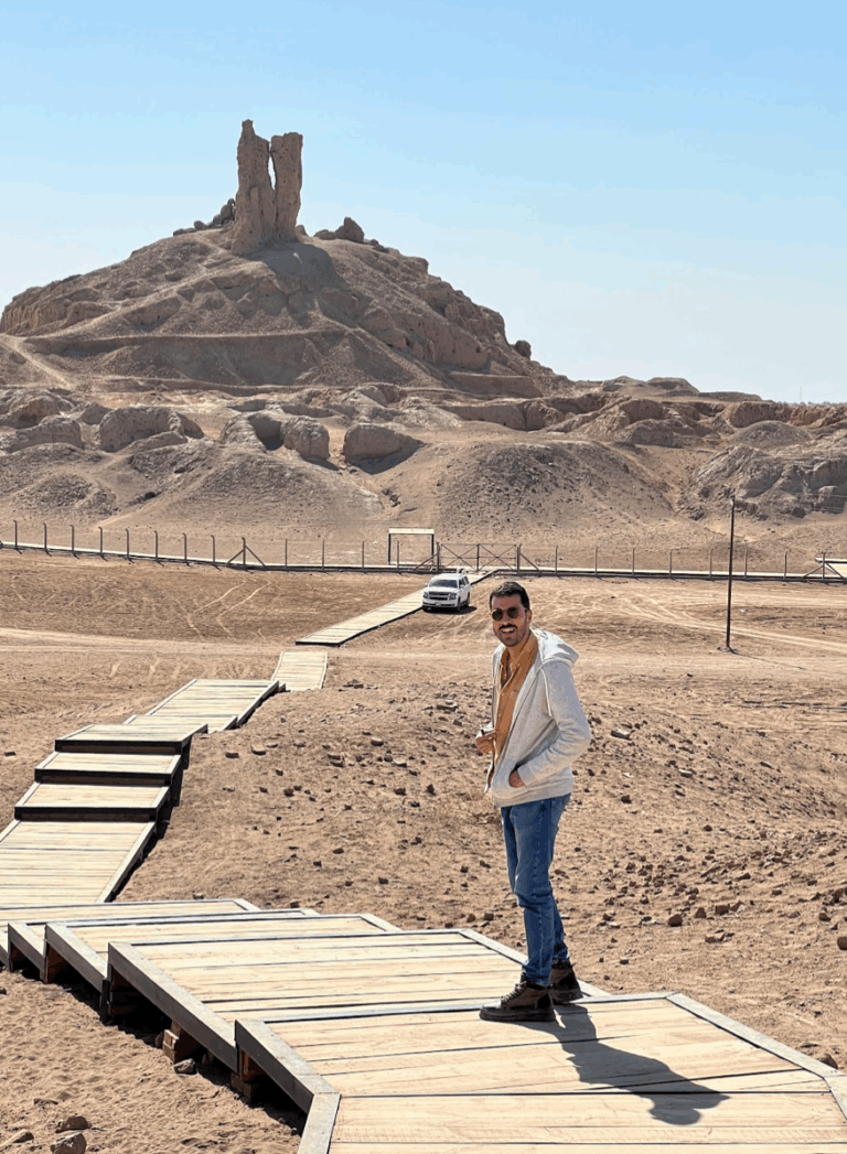 A man stands on a wooden walkway leading to a mound of ruins, with a car parked nearby under a clear sky.
