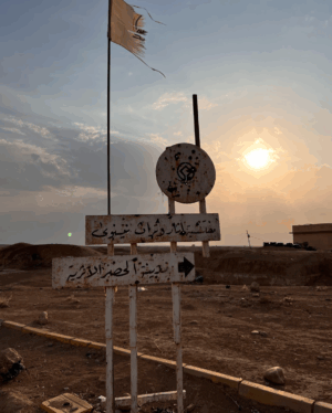Day Tour Mosul to Hatra A weathered sign in a desolate landscape with Arabic writing points the way, a tattered flag waving above under a setting sun.